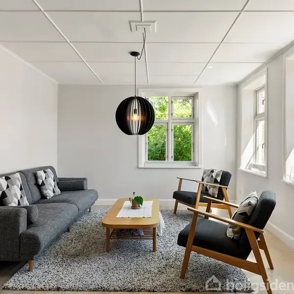 En stue med en grå sofa og to stole står mod et tæppebelagt gulv. Et sofabord er centreret, mens en sort loftslampe hænger over. Store vinduer lader dagslys ind.