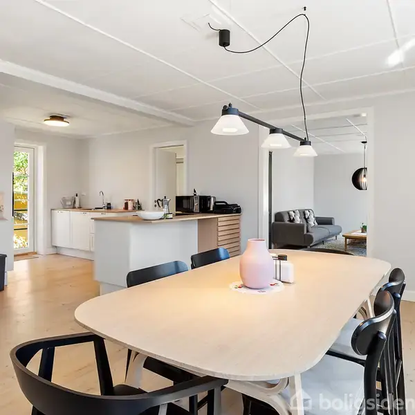 Et træbord med stole står i et lyst, moderne køkken-alrum med åben adgang til en stue. Et loftlys hænger over bordet, mens en sofa ses i baggrunden.
