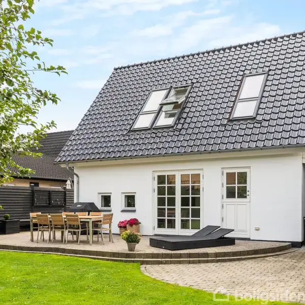 Et hvidt hus med sort tag står på en grøn græsplæne. Foran er et terrasseområde med et bord og stole, omgivet af hæk og træer.