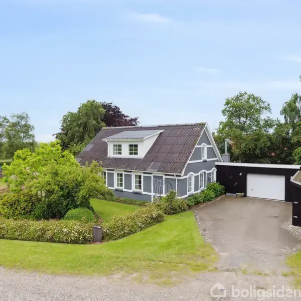 Et gråmalet hus med tegltag står i en frodig have med buske og træer. Der er en indkørsel til en garage og omgivelserne er landlige med græsplæner og grusvej.