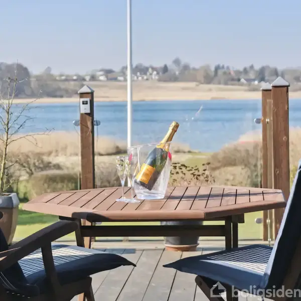 Et rundt bord med en flaske champagne i en isspand står på en terrasse. To stole omkranser bordet. I baggrunden ses en sø med omgivelser af græs og træer.