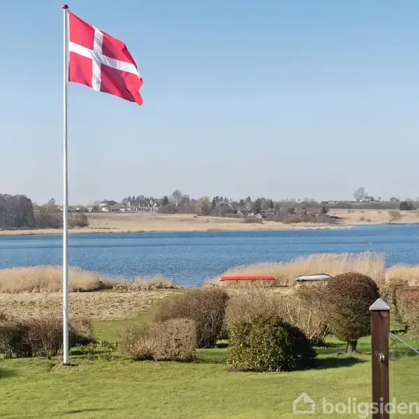 Det danske flag vajer på en flagstang i en velplejet have. Baggrunden viser en sø og en landlig horisont med skove og marker under en klar, blå himmel.