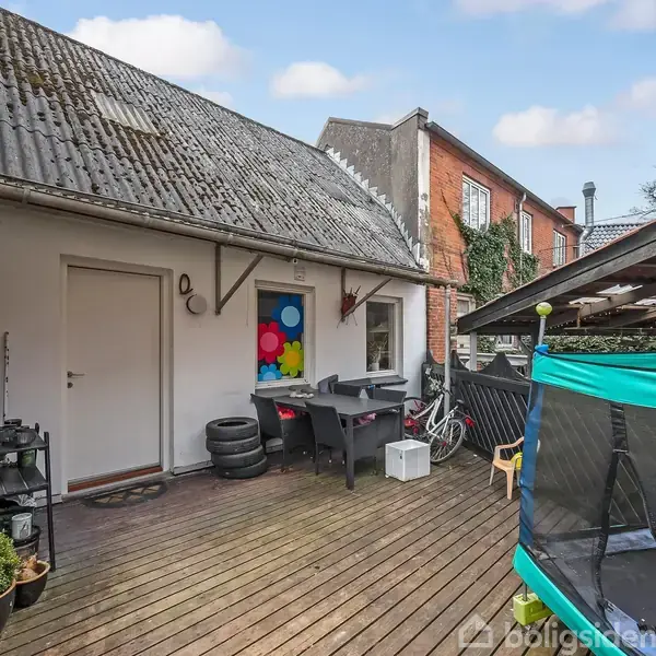 En terrasse med træbrædder, har et bord og stole. En lille trampolin står til højre. Baghaven er omgivet af murstensbygninger. Farverige blomsterdekorationer hænger på en hvid væg.