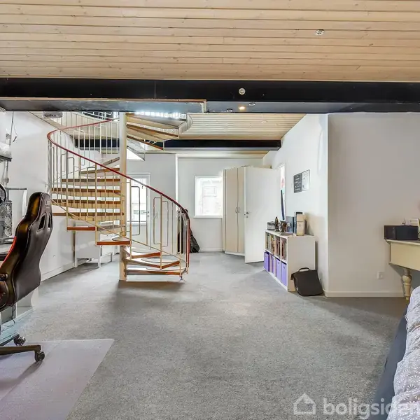 Et kontorområde med en kontorstol og computerbord står ved siden af en vindeltrappe. I baggrunden ses en seng, en kommode og en åben dør i et lyst rum.