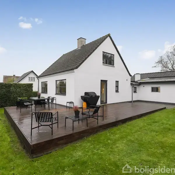 Hus med hvid facade og sort tag står på en træterrasse med havemøbler og grill. Omgivet af grøn græsplæne, med hegn og træer i baggrunden under en klar himmel.