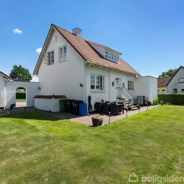 Hvidt hus med rødt tag står i solrig have, omgivet af grøn plæne og hække. Der er havemøbler på flisebelagt terrasse. Tydelig blå himmel og andre huse i baggrunden.
