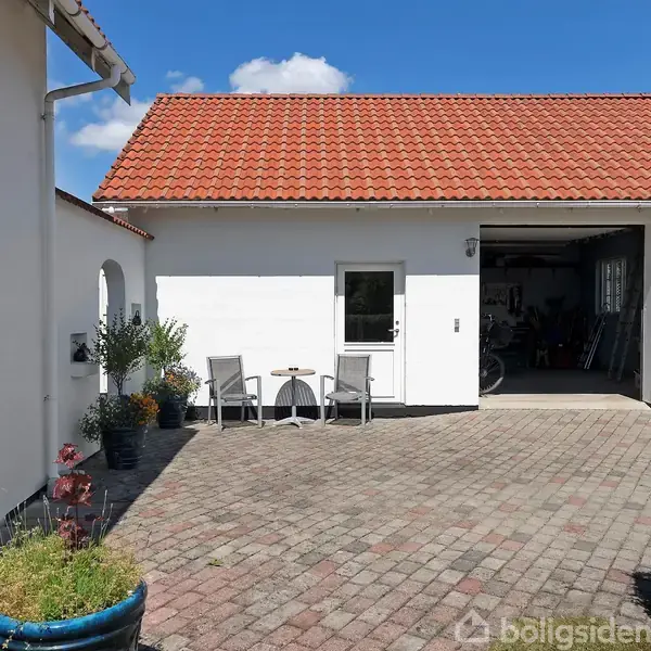Et hvidt garagebygning med rødt tegltag står i solen. Foran er en flisebelagt gårdsplads med to grå stole og et lille bord. Skyfri blå himmel i baggrunden.
