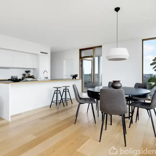 En moderne spisestue med rundt bord og seks stole i et lyst rum. Et stort vindue giver udsigt over byen. Et køkkenområde med barstole er i baggrunden.