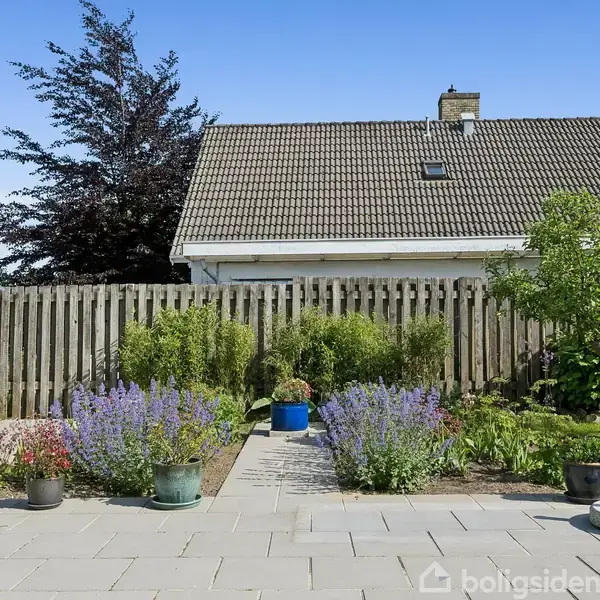 En velplejet have rummer blomstrende planter i krukker, omkranset af et hegn foran et hus med skråtag. Til højre ses et træ ved siden af en velholdt græsplæne.