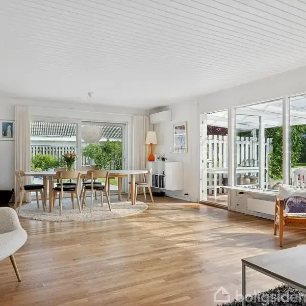 Hus med trekantet tag står i en velholdt have med græs og terrasse. Naboer ses i baggrunden, og klare blå himmel danner kulissen.