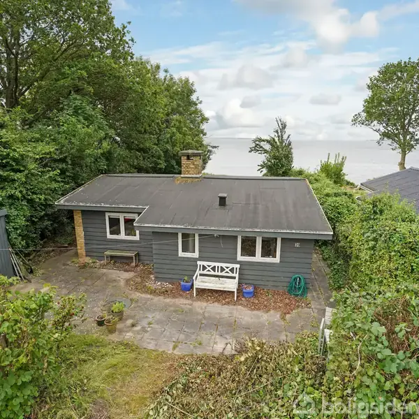 Et gråt sommerhus med sort tag ligger omgivet af frodig grøn vegetation. Foran huset er der en flisebelagt terrasse. I baggrunden ses havet og flere træer.