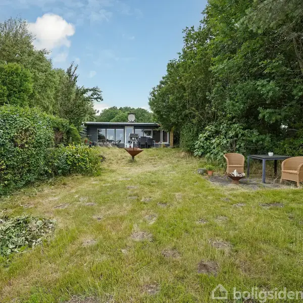 Et sommerhus står i baggrunden omgivet af frodig grøn bevoksning. Foran ses en græsplæne med et havebord og to kurvestole. Himlen er delvist skyet.