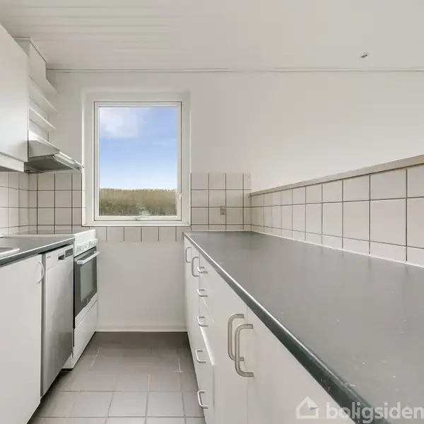 Køkken med hvidt interiør og grå bordplader. Køleskab og ovn til venstre. Et vindue med udsigt til trætoppene skaber lys. Flisebelægning giver et rent udtryk.