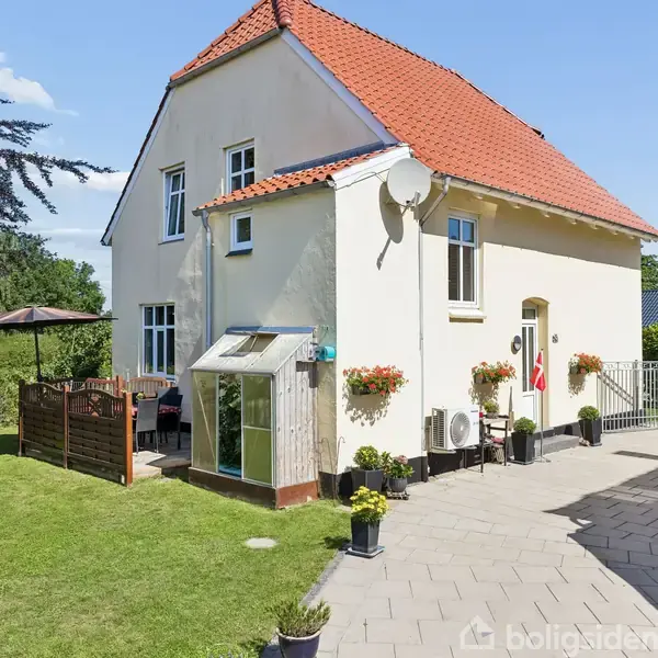 Et hvidt to-etagers hus står i solskin med rødt tegltag og blomsterkrukker ved indgangen. Ved siden af huset er der en terrasse med havemøbler og omkransende hæk.