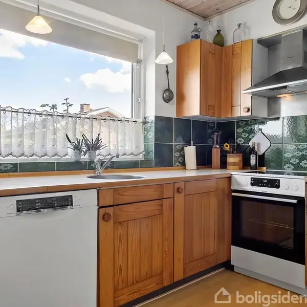 Køkken med træskabe og opvaskemaskine. Ovn og emhætte integreret i skabene. Mørkegrønne fliser på væggen. Et vindue med udsigt til en blå himmel og naboens tag.