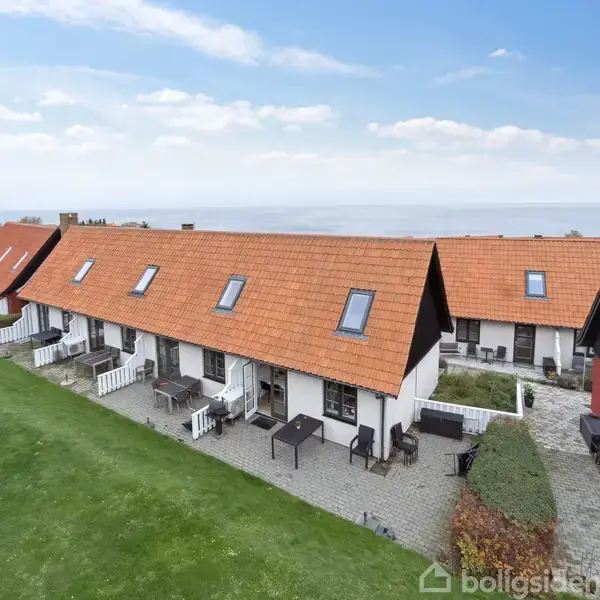 Rækkehuse med røde tegltage står side om side, omgivet af grønne plæner. Små terrasser strækker sig fra hver enhed, og i baggrunden kan havet anes under den blå himmel.