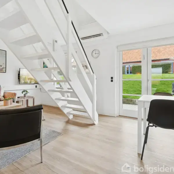 En moderne stue med hvide trapper fører opad, omgivet af møbler og vægkunst. Franske døre fører ud til en græsplæne. Gulv af lyst træ.