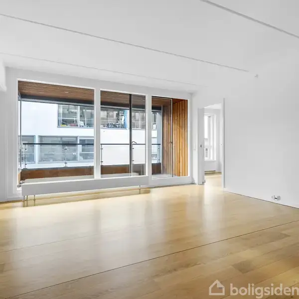 Stue med trægulv, store glasdøre, der åbner ud til en altan; moderne, lyse omgivelser med hvidmalede vægge, åbent køkkenhjørne og udsigt til nabobygninger.