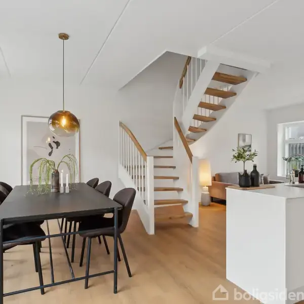 En moderne spisestue har et sort spisebord med seks stole. Rummet er lyst med hvide vægge, trappe til første sal og åben forbindelse til køkkenet. En gul lampe hænger over bordet.