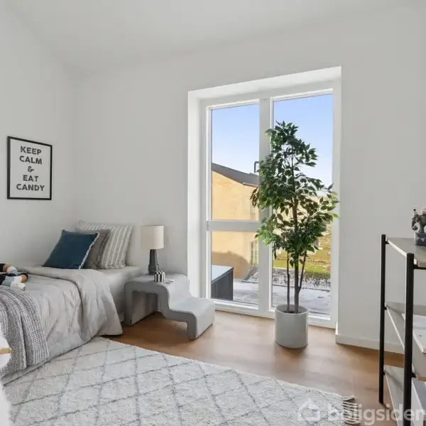 En enkeltseng står i et lyst værelse med en plakat på væggen med teksten "KEEP CALM & EAT CANDY." Et stort vindue giver udsigt til en terrasse. En plante og hylder med pynt supplerer rummet.