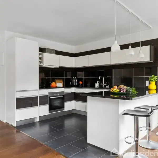 Et moderne køkken med hvide og sorte elementer. Tre barstole står ved en hvid køkkenø med planter og frugt. Elegant lys hænger fra loftet, og gulvet er mørkt flisebelagt.
