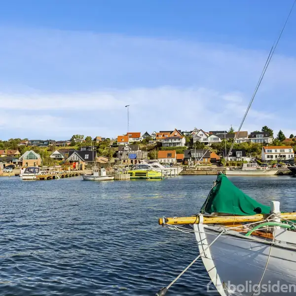 En sejlbåd ligger for anker i en rolig havn, omgivet af farverige huse på en kystlinje under en klar blå himmel.