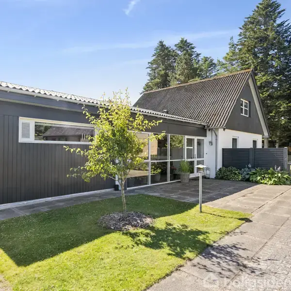 Hus i moderne stil med sort-hvid facade står på en velplejet græsplæne. En indkørsel fører mod hovedindgangen, omgivet af en have med træer og buske, beliggende i et skovområde.