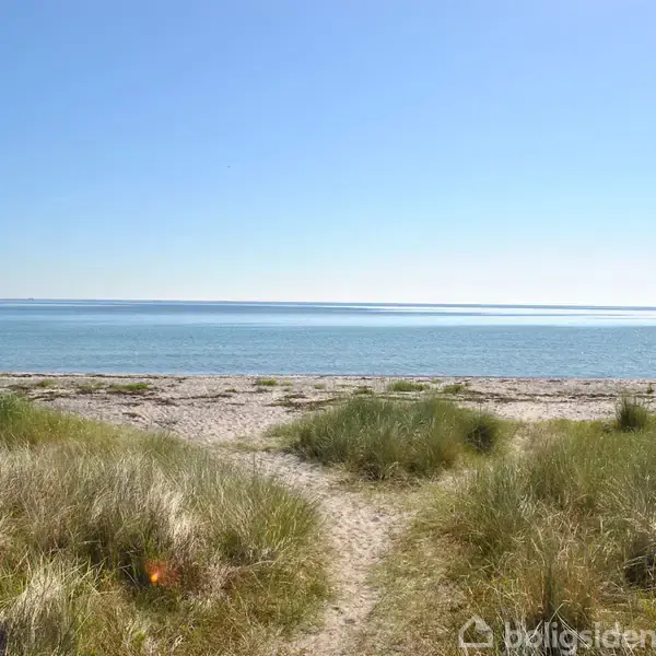 Sti løber gennem græsbevokset klit mod fredelig sandstrand og stille, blå hav under en klar, solrig himmel.