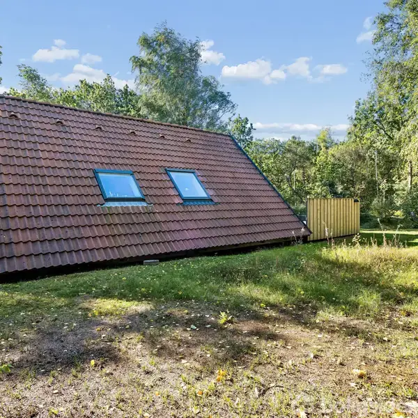 Et skråt hus med et rødt tegltag og to vinduer står stille midt i en grøn skovlysning under en klar blå himmel.