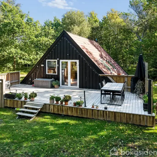 A-formet sort hus står stille på en stor træterrasse med planter i potter og havemøbler. Det er omgivet af en grøn græsplæne og tæt skov bagved.