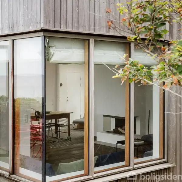 Glasparti af moderne hus viser interiør med spisebord og stole. Udenfor ses træer og natur. Indvendigt er der en pejs og lyse vægge, der skaber en hyggelig atmosfære.