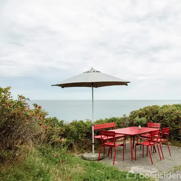 Røde havemøbler med en parasol står stille på en terrasse med udsigt til havet. Omgivet af grønne buske og blomster under en overskyet himmel.