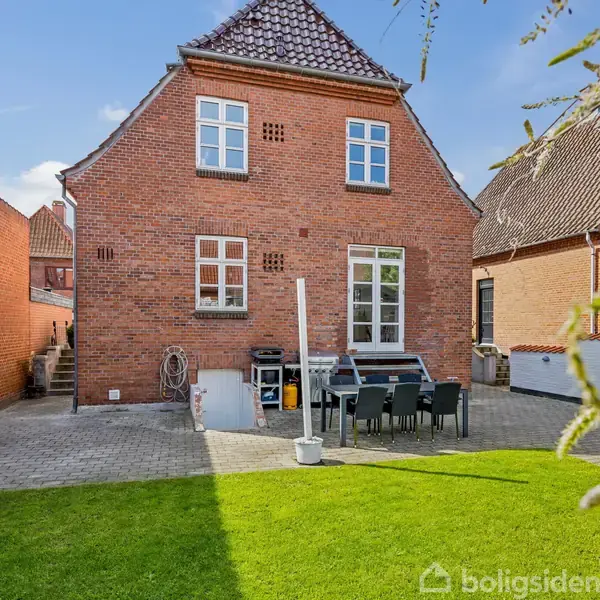 Et rødt murstenshus står ved en brolagt terrasse med et bord og stole. Græsplæne omkranser terrassen, mens en mur forbinder huset med en anden bygning.