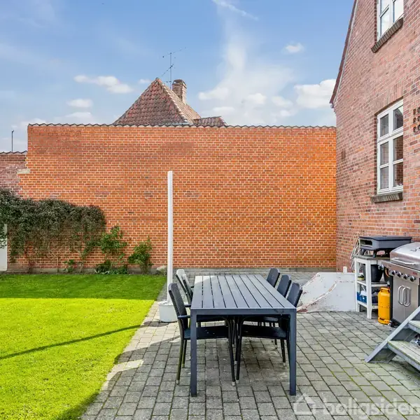 Et sort havebord med seks stole står på en flisebelagt terrasse. Ved siden af er en grill. Omkring er en murstensvæg og et græsareal foran en villa.