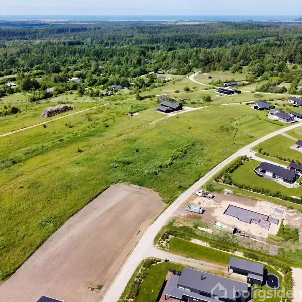 Luftfoto over parcelhusområde. Huse er spredt ud over grønne græsarealer med omgivende skovområde. Flere grusveje forbinder de enkelte ejendomme gennem landskabet.