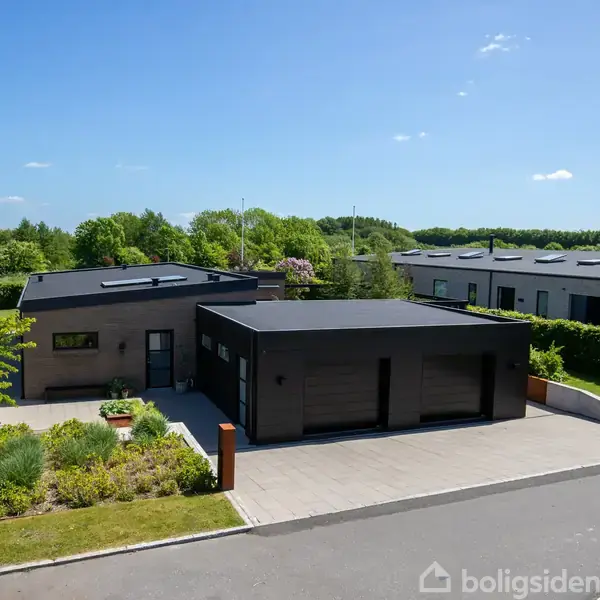 En moderne villa med fladt tag står på en velplejet grund med grønne buske. En flisebelagt indkørsel fører til en dobbelgarage. Baggrunden viser flere huse og grøn natur.