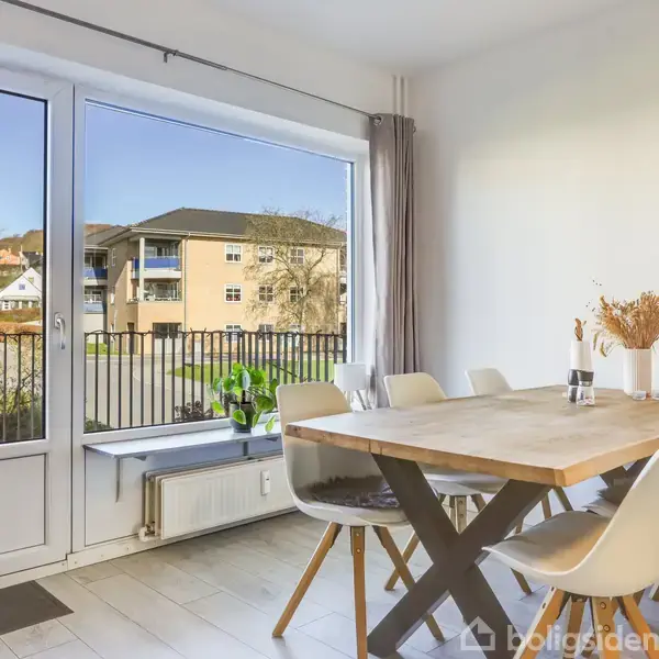 Spisebord med seks stole står i et lyst rum. En glasdør med udsigt til en boligblok udenfor. Gardiner hænger ved siden af døren, og en vase står på bordet.