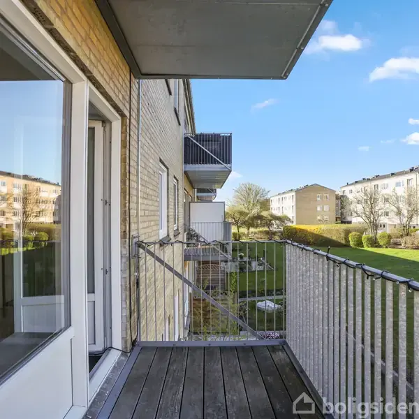 Balkon med trægulv strækker sig ud fra en lejlighed. Hyggelig udsigt til grønne områder og nabobygninger. Klar blå himmel skaber en lys og indbydende atmosfære.