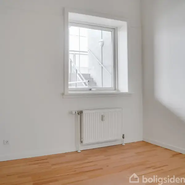 Et enkelt, tomt rum med lyst trægulv har en radiator under et vindue. Uden for vinduet ses en betontrappe. Væggene er hvide, og der er en døråbning til højre.