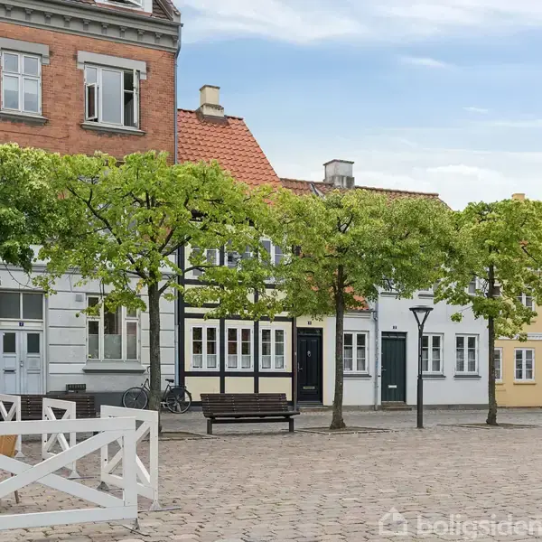 Torv med brosten og træer danner ramme om bygninger med farverige facader. En bænk og gadelygte ses i forgrunden, mens himlen er let overskyet.