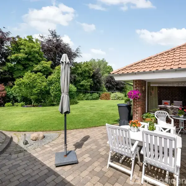 Terrassemøbler står på en brolagt terrasse med en lukket parasol. Blomster pryder bordet, omgivet af grønne græsplæner og buske. Solrigt vejr og blå himmel med lette skyer.