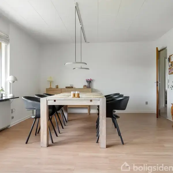Langt spisebord med sorte stole står i et lyst spiseområde. Der er store vinduer til venstre, som lukker naturligt lys ind, og lyse møbler omgiver rummet.