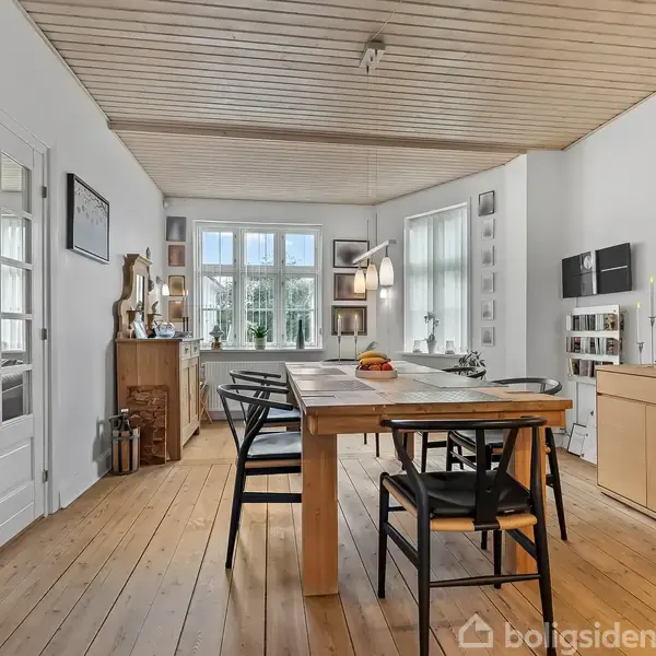 Spisebord med sorte stole står i et lyst rum med trægulv og træloft. Store vinduer kaster naturligt lys ind, omgivet af planter og dekorative genstande. Maleri på væggen.