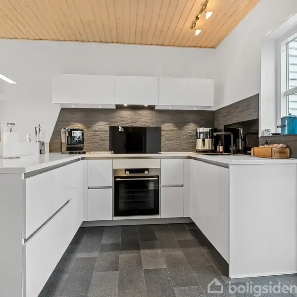 Køkken med hvide skabe og skuffer omkring indbygget ovn, kaffemaskine og skærebræt. LED-lys på trælignende loft og flisegulv. Vindue og dør til højre, moderne og minimalistisk design.