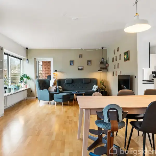 En murstensvilla står på en grøn plæne med en terrasse foran. Et drivhus er placeret ved siden af huset. Træer omgiver området under en klar blå himmel.