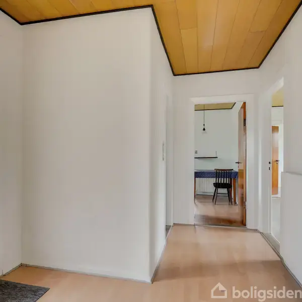 En entré med hvide vægge og trægulv fører til en lys gang. En åben dør viser et rum med blå stol. Hvid dør til venstre, radiator til højre.