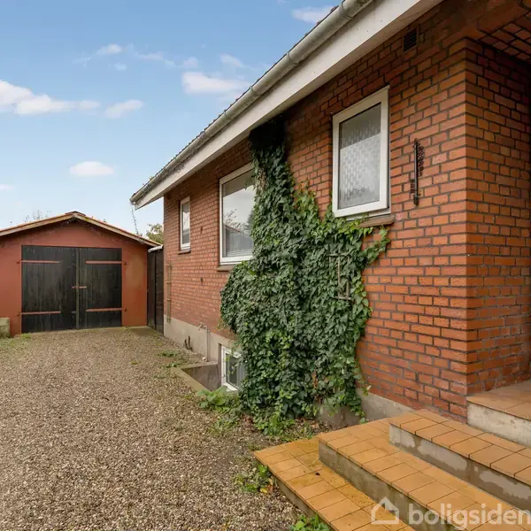 Murstenshus med efeu vokser op ad facaden, beliggende ved en grussti. Huset har hvide vinduer og en brun trædør. I baggrunden ses en rød garage med sorte døre.