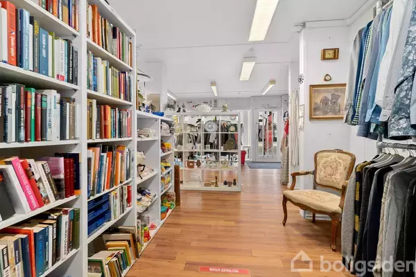 Bøgerne står på hylden i en veloplyst butik, omgivet af antikt inventar og tøj. En vintage stol er placeret ved siden af hylden, med trægulv under.