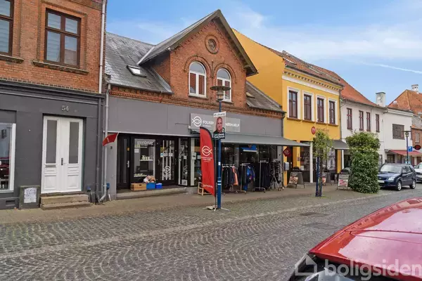 Gade med brosten og huse i forskellige farver. Butik med tøjstativ og skilt med teksten "Folkekirkens Nødhjælp". Bil parkeret på vejen under en blå himmel.