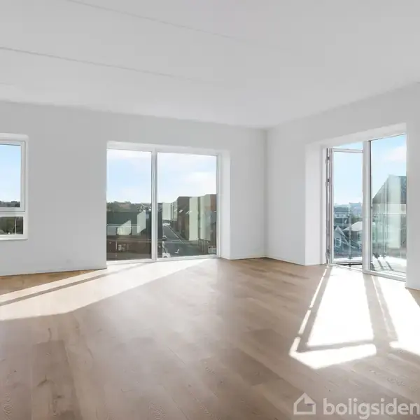 Et lyst og rummeligt værelse med trægulv, store vinduer og altandøre, der åbner ud til en balkon med udsigt over en by. Sollys strømmer ind og oplyser rummet.